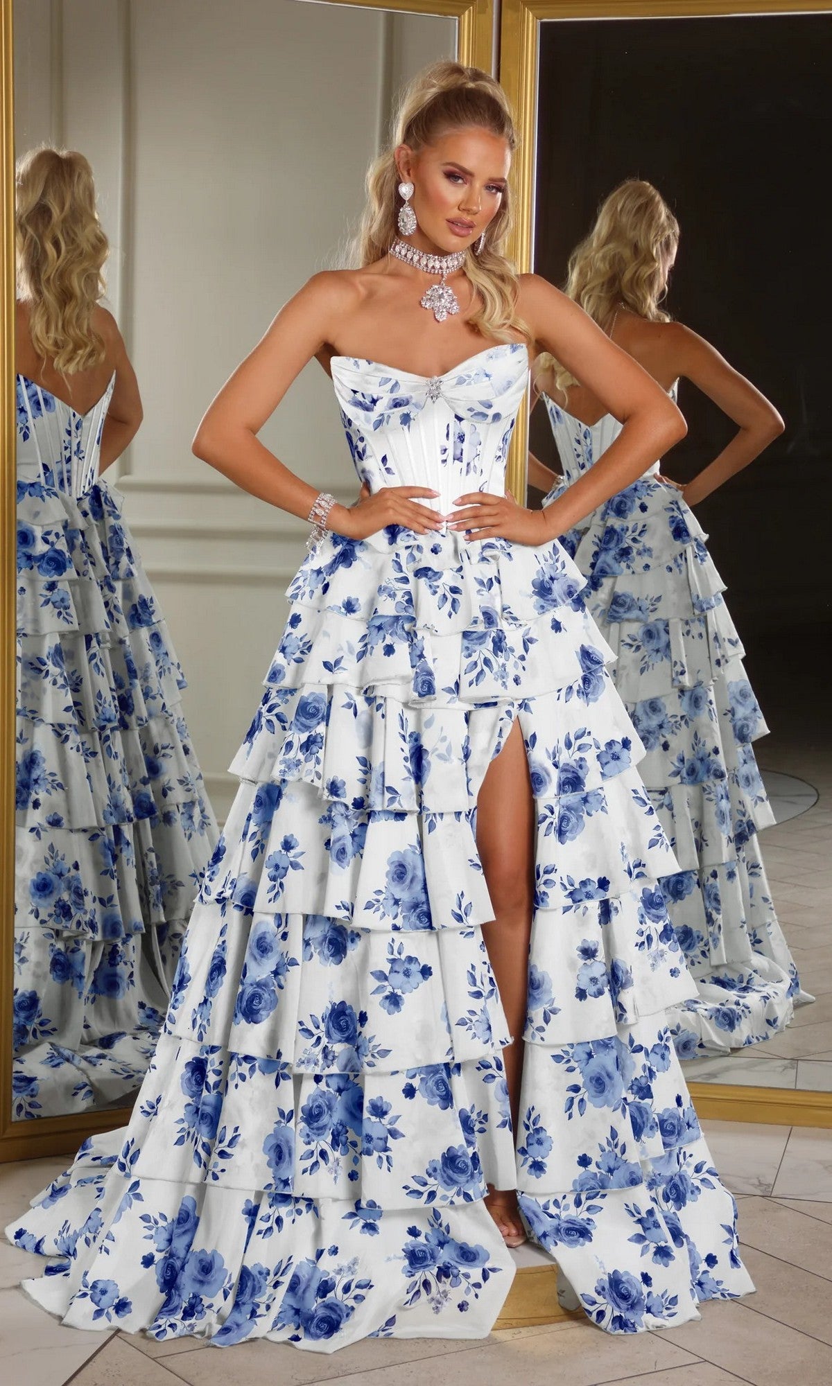 A woman in the Portia and Scarlett PS25966 floral ball gown, featuring a strapless white/blue corset bodice, ruffled tiered skirt, thigh-high slit, and jewelry, stands before mirrors.